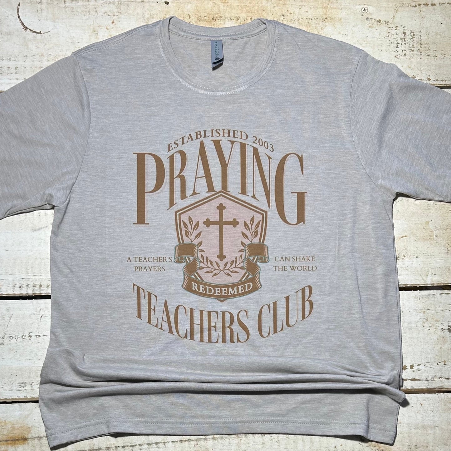 Alt Text: A T-shirt from the Red Oak Company's Faith Collection, featuring the "Praying Teacher Club" design. The graphic showcases a teacher in prayer, symbolizing unity, strength, and devotion. The shirt is made from a comfortable Next Level poly/cotton blend, perfect for expressing both faith and style.