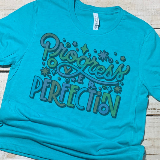 **Alt Text for "Progress Over Perfection" Teacher Shirt Image:**  A teacher stands in a classroom, wearing a soft Heather Sea Green poly/cotton shirt with the words "Progress Over Perfection" printed in vibrant, cheerful colors. The teacher has a warm smile and is surrounded by eager students, all engaged in learning activities. This teacher shirt embodies positivity and inspiration for educators of all grades.