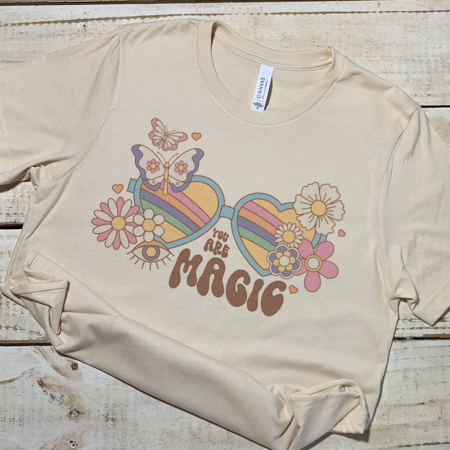 Alt Text: A soft cream Bella-Canvas teacher shirt with the words "You Are Magic" printed in groovy typography. The shirt is laid out against a neutral backdrop, highlighting its comfortable and versatile design.