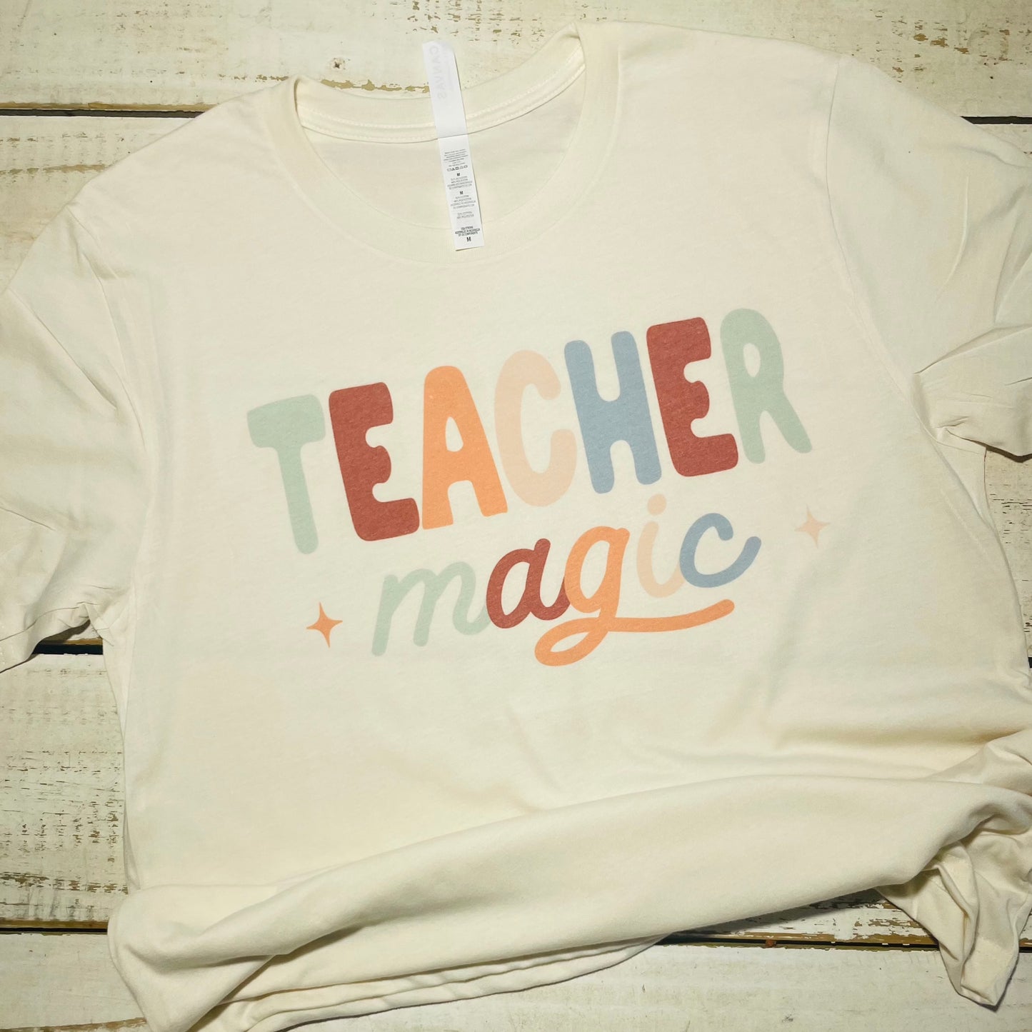 **Alt Text for "Teacher Magic" Shirt Image**  "An attractive teacher wearing the 'Teacher Magic' shirt, featuring a whimsical design with a magician's hat, wand, and spellbook. The shirt is made of a comfortable poly/cotton blend and comes in vibrant colors. The teacher smiles while standing in a classroom filled with books and educational materials."