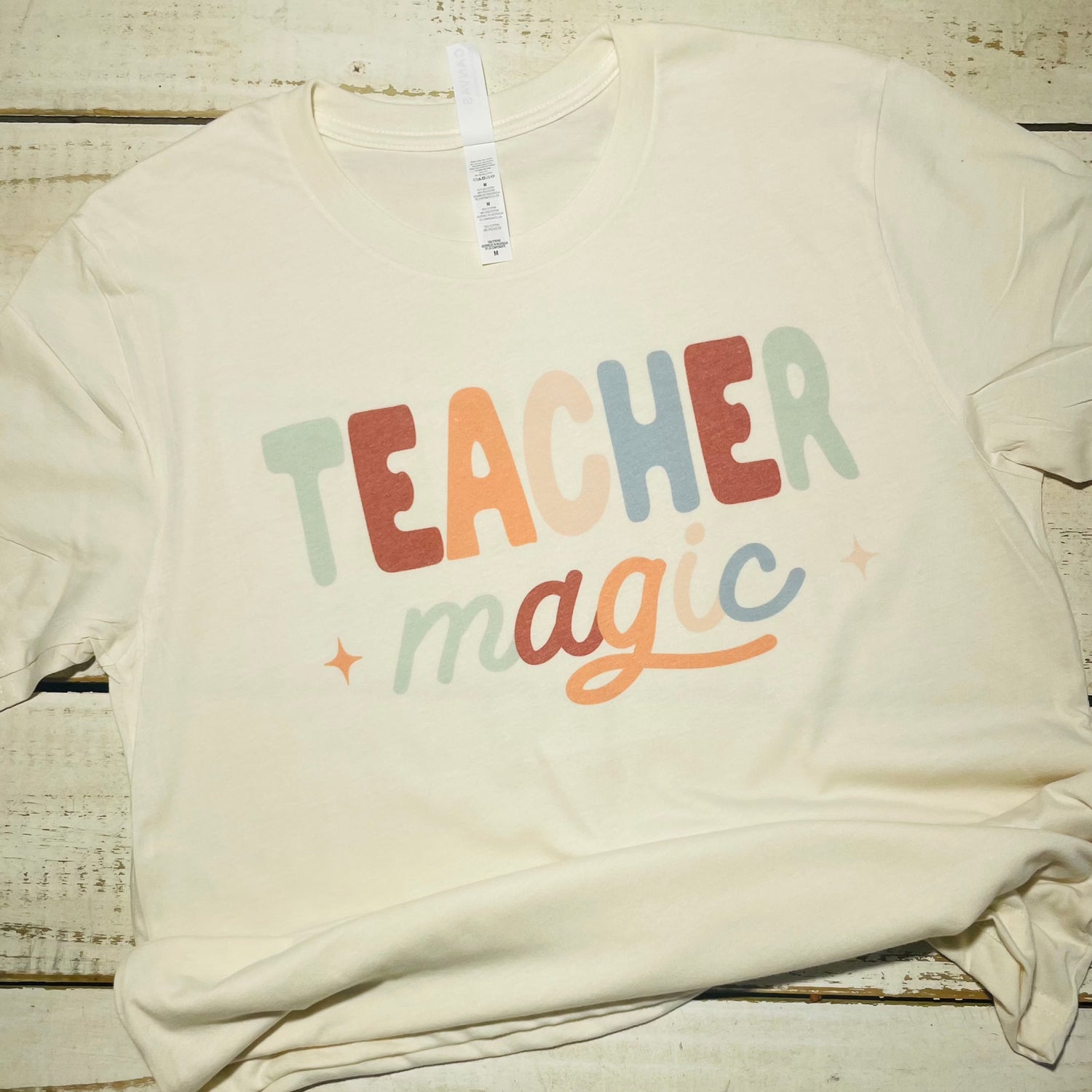 **Alt Text for "Teacher Magic" Shirt Image**  "An attractive teacher wearing the 'Teacher Magic' shirt, featuring a whimsical design with a magician's hat, wand, and spellbook. The shirt is made of a comfortable poly/cotton blend and comes in vibrant colors. The teacher smiles while standing in a classroom filled with books and educational materials."