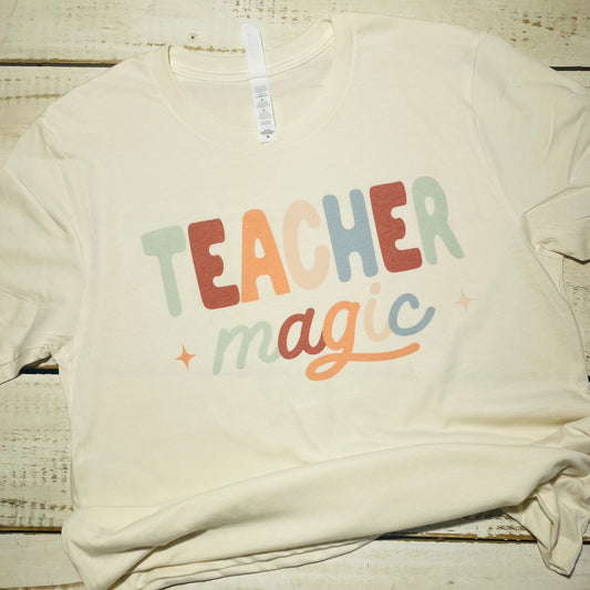 **Alt Text for "Teacher Magic" Shirt Image**  "An attractive teacher wearing the 'Teacher Magic' shirt, featuring a whimsical design with a magician's hat, wand, and spellbook. The shirt is made of a comfortable poly/cotton blend and comes in vibrant colors. The teacher smiles while standing in a classroom filled with books and educational materials."