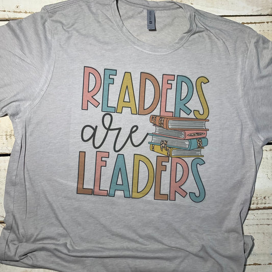 "Vibrant Next Level Silver teacher shirt featuring the empowering message 'Readers are Leaders.' The shirt is elegantly displayed against a backdrop of books, symbolizing the educator's role in nurturing knowledge and curiosity. The design captures the essence of teaching with style and purpose, making it a must-have addition to any teacher's wardrobe."