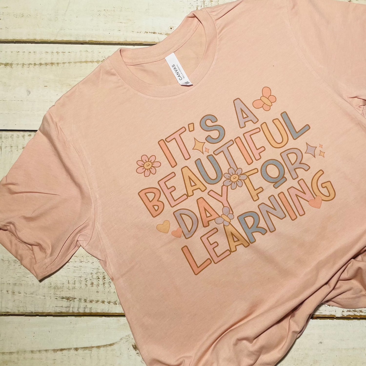 **Alt Text for "It's a Beautiful Day For Learning" Teacher Shirt:**  A teacher shirt with the text "It's a Beautiful Day For Learning" in a playful yet elegant font. The shirt is in Heather Peach color and is made from soft Bella-Canvas fabric. The design embodies positivity and dedication to education, making it a perfect choice for teachers of all ages. The shirt is versatile for both classroom and casual wear, encouraging a love for learning wherever it's worn.