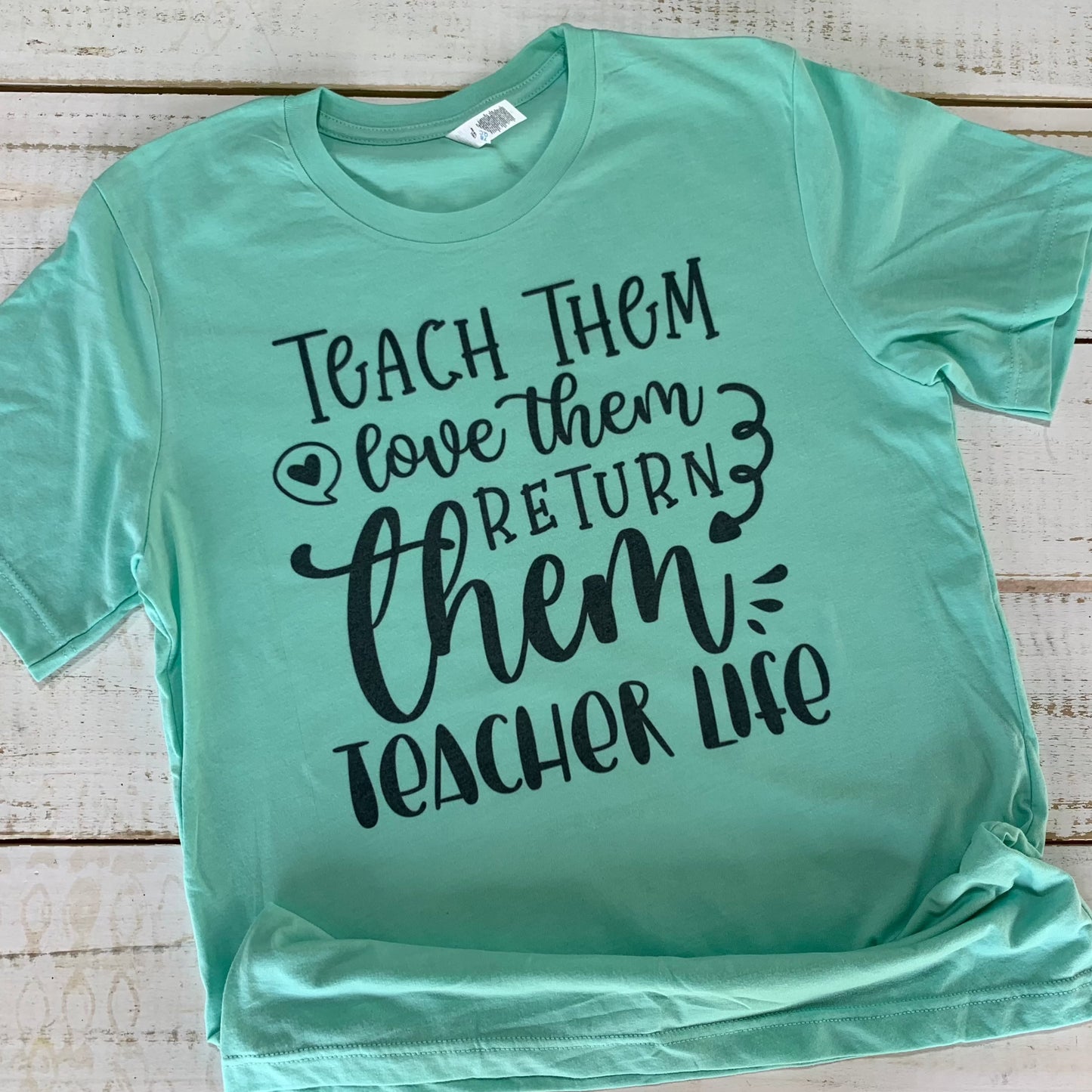 "Teacher shirt with empowering quote: 'Teach Them Love Them Return Them Teacher Life' on a Bella-Canvas Heather Mint background."